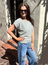 A woman wearing a light sage green short-sleeve knit top and blue jeans stands outdoors against a stucco wall with shadows.