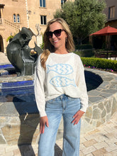 Woman wearing a white sweater with blue fish design and jeans standing in front of a fountain.