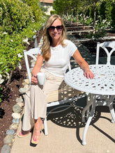 Woman sitting at a white outdoor table holding a cup, surrounded by greenery.