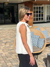 Woman in a white sleeveless top and sunglasses standing next to a wicker chair with patterned cushions.