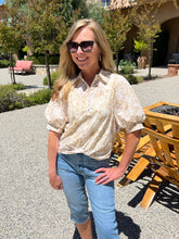 Woman wearing sunglasses and a patterned blouse standing outdoors with greenery and a building in the background.