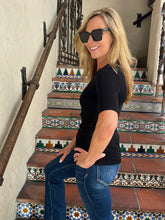 A woman with blonde hair wearing a black short-sleeve t-shirt and blue jeans stands on a tiled staircase, looking back over her shoulder. She is wearing black sunglasses.