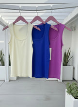 Three tank tops in white, blue, and purple hanging on pink hangers against a white wall.