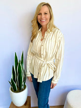 Woman wearing a striped shirt and blue jeans standing next to a potted plant against a light blue wall.