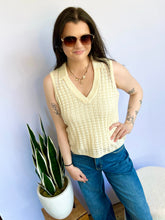 Woman wearing a cream sleeveless top and blue jeans standing next to a potted plant against a white wall.