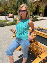 Woman in a turquoise sleeveless top and blue jeans standing outdoors near wooden benches.