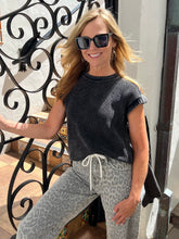 A woman wearing a dark gray ribbed short-sleeve knit top and light gray leopard-print pants, standing outdoors next to a black wrought-iron gate.