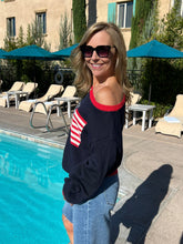 Woman standing by a poolside wearing sunglasses and a navy sweater with red and white accents.