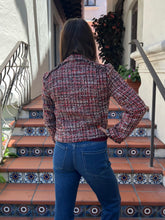 A woman viewed from behind wearing a red, white, and blue tweed jacket and blue jeans, standing on outdoor tiled stairs.