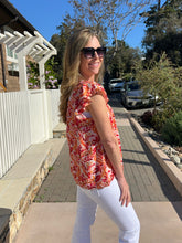 Woman in a floral top and sunglasses standing on a sidewalk with a house and cars in the background.