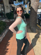 Woman in a mint tank top and black pants standing on a sidewalk with trees and buildings in the background.