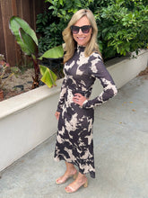 A woman modeling a long-sleeve midi dress with a dark gray and beige abstract pattern, standing outdoors on a concrete walkway next to a planter with green foliage.