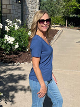 Woman in a blue shirt and sunglasses standing on a sidewalk with greenery in the background