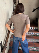 A woman wearing a fitted taupe short-sleeve knit top and blue jeans stands on a tiled staircase indoors. She is also wearing dark sunglasses and a delicate necklace.