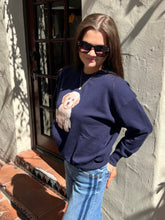 A woman wearing a navy blue crewneck sweatshirt with a cream-colored dog graphic on the front, paired with light blue jeans, standing outdoors against a stucco wall.