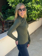 A woman wearing an olive green long-sleeve mock neck top and dark sunglasses, posing outdoors on a concrete walkway with green plants and a beige wall in the background.