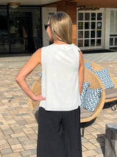 Woman in a white sleeveless top and black pants standing outdoors near a patio chair with blue and white pillows.