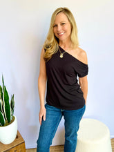 Woman wearing a black one-shoulder top and blue jeans standing against a white wall with a plant in the background.
