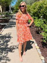 Woman in an orange floral dress standing on a sidewalk with greenery in the background