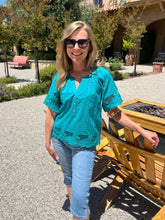Woman in a turquoise blouse and sunglasses standing outdoors near a wooden structure.