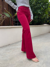 A person wearing bright magenta flare jeans with a light gray long-sleeve top and beige slide sandals, standing outdoors on a concrete patio with green plants and a low wall in the background.