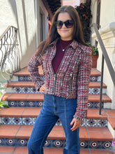 A woman wearing a red and black tweed zip-up jacket over a burgundy top and blue jeans, standing on tiled outdoor stairs.