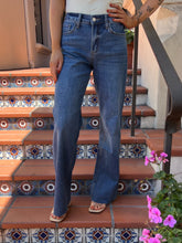 A person wearing blue denim jeans stands on outdoor tiled stairs, with the jeans shown from the waist down. The setting includes terracotta steps, decorative blue-and-white tile bands, a black metal railing, and pink flowers in the foreground.