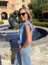 Woman in a blue sleeveless top and jeans standing in front of a fountain with a building and garden in the background.