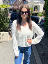 A woman wearing a white cable-knit long-sleeve sweater and blue jeans stands outdoors on a residential street, wearing dark sunglasses.