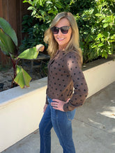 A woman wearing a brown long-sleeve top with black polka dots and blue jeans, standing outdoors in front of green plants and a beige wall.