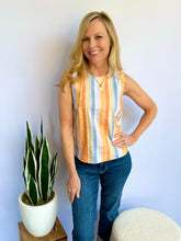 Woman wearing a striped sleeveless top and jeans standing in a room with a plant and ottoman.
