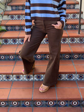 A person wearing brown wide-leg pants and a blue and brown striped sweater, standing on terracotta tiled stairs with decorative blue and white patterned risers.