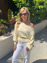 Woman wearing a yellow off-shoulder top and white pants standing outdoors with greenery in the background.