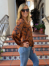 A woman wearing a rust-orange long-sleeve blouse with a dark abstract floral pattern and light blue jeans, standing on outdoor tiled steps.