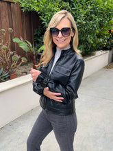 A woman wearing a black faux leather moto jacket, dark sunglasses, and gray jeans, standing outdoors on a concrete patio with green plants and a wooden fence in the background.