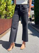 A person outdoors wearing black wide-leg cropped pants with a white top and beige heeled sandals, standing on a paved walkway with greenery and a building in the background.