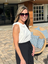 Woman wearing sunglasses and a white top with black pants standing outdoors near a wicker chair with blue and white cushions.