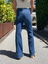 A person standing outdoors on a paved path wearing blue high-waisted flare jeans, a cream sleeveless top, and tan heeled sandals, viewed from behind.