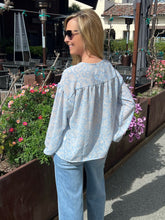 Woman wearing a light blue floral blouse and jeans standing outdoors with flowers and chairs in the background.