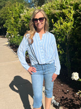 Woman wearing a light blue striped shirt and jeans standing outdoors with greenery in the background