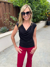 A woman wearing a black sleeveless cowl-neck top and bright red pants, standing outdoors on a concrete patio with a garden fence and plants in the background.