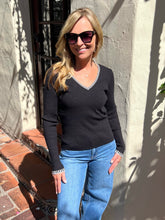 A woman wearing a black long-sleeve V-neck sweater with white patterned trim on the collar and cuffs, paired with blue jeans, standing outdoors against a stucco wall.