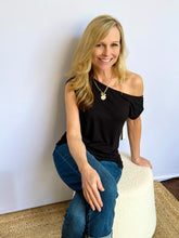 Woman sitting on a white stool against a plain wall wearing a black top and blue jeans.