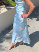 Woman wearing a light blue floral dress standing on a concrete surface with plants in the background.