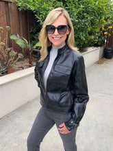 A woman wearing a black leather jacket, gray turtleneck, and dark sunglasses stands outdoors on a concrete walkway with green plants and a wooden fence in the background.