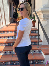 A woman wearing a white short-sleeve crewneck t-shirt and dark jeans stands on outdoor tiled steps, looking over her shoulder. She is wearing black sunglasses and hoop earrings.