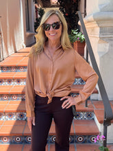 A woman wearing a tan long-sleeve button-up shirt tied at the waist and dark brown pants, standing on tiled outdoor stairs.