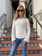 A woman wearing a cream ribbed long-sleeve top and blue jeans stands on tiled outdoor stairs in front of a building.