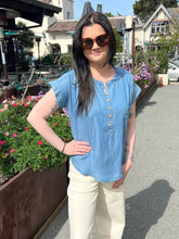 Woman wearing a chambray henley top and sunglasses standing outdoors with plants and buildings in the background.