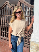 A woman wearing a beige short-sleeve tie-front top and blue jeans stands outdoors in front of a decorative brown metal railing.